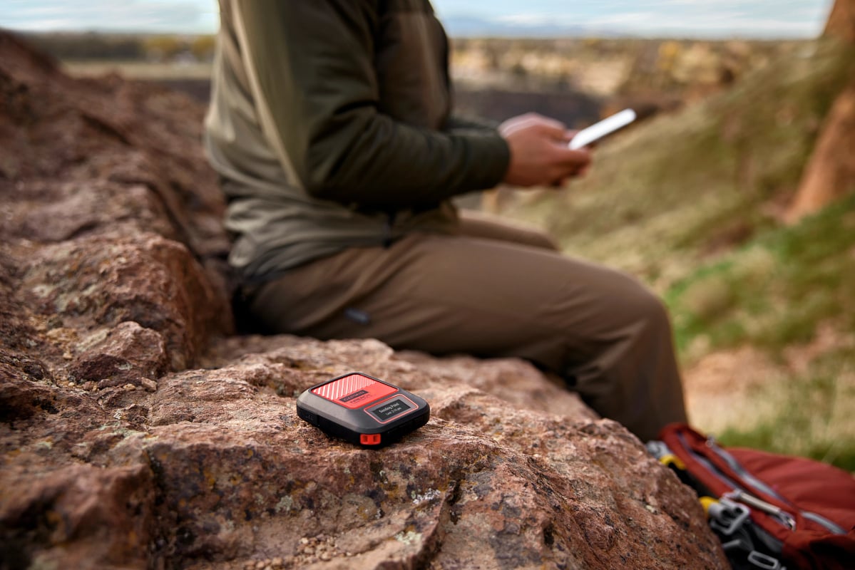 A man in a green shirt and brown pants uses a phone while the Garmin inReach Messenger Plus satellite communicator is on a rock.