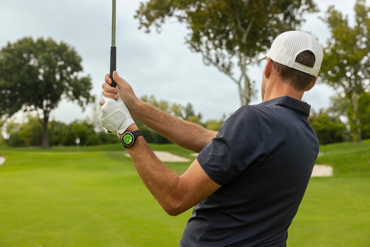 A man in a navy blue polo and white hat finishes swinging a golf club.
