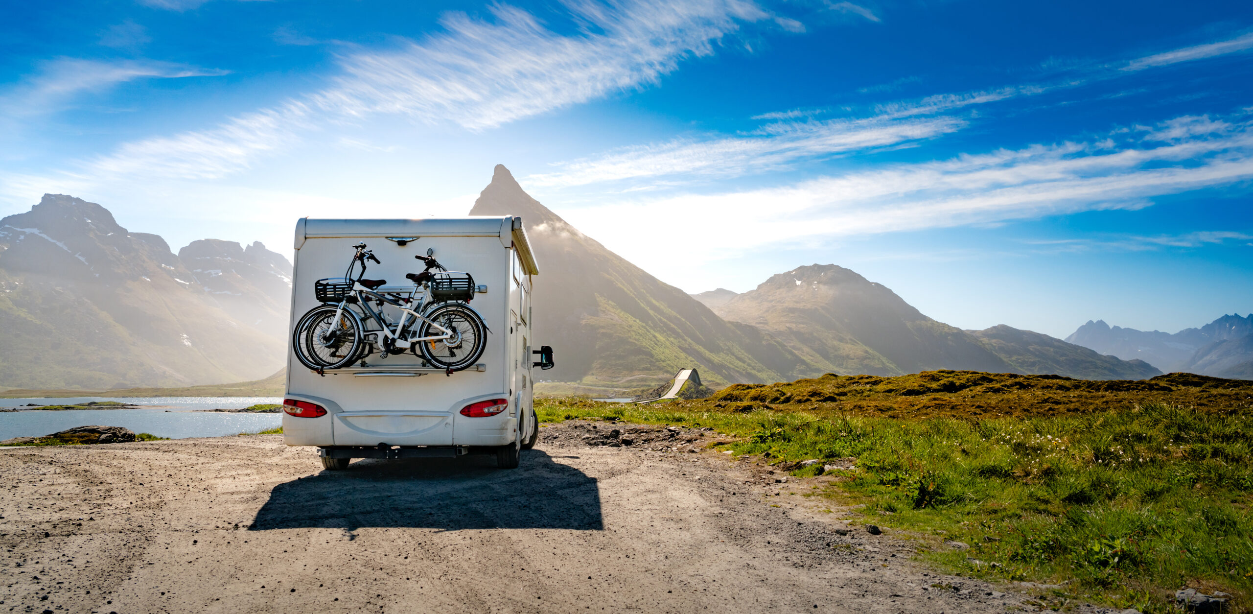 Back view of an RV on a dirt road with a bike attached to the back. Behind the RV lies a mountain range.