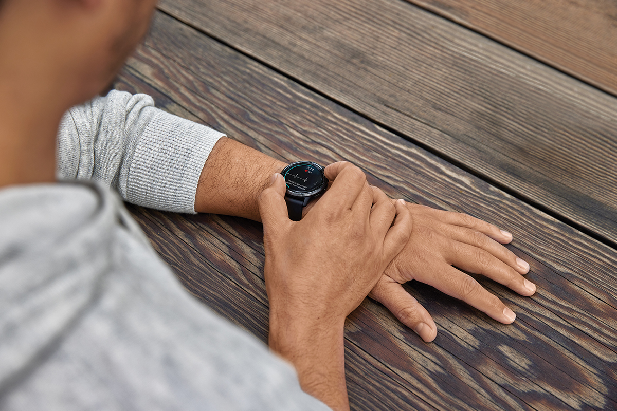 A man in a gray sweatshirt uses the Garmin ECG app on his Garmin smartwatch. The watch screen says, "The ECG app cannot detect heart attacks."