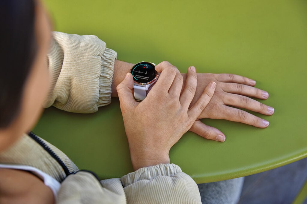 A woman in a tan jacket uses the Garmin ECG app on her Garmin smartwatch. The watch screen says, "The ECG app cannot detect heart attacks."