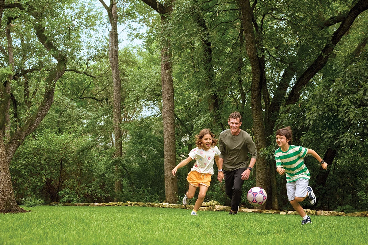 Two children wearing Garmin Bounce 2 smartwatches play with a soccer ball with their father in an open grassy area.
