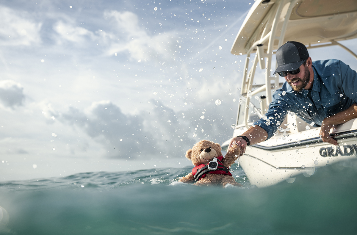 A man reaching into the water from his boat grabbing a teddy bear out of the water. The teddy bear has an MOB tag and life jacket on.